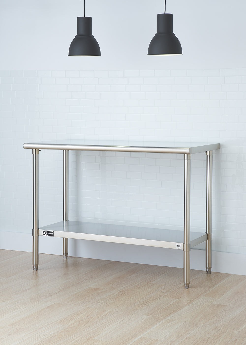 A stainless steel utility table with an undershelf stands against a white brick wall. Two black pendant lights hang from the ceiling above the table.