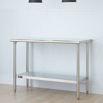 A stainless steel utility table with an undershelf stands against a white brick wall. Two black pendant lights hang from the ceiling above the table.
