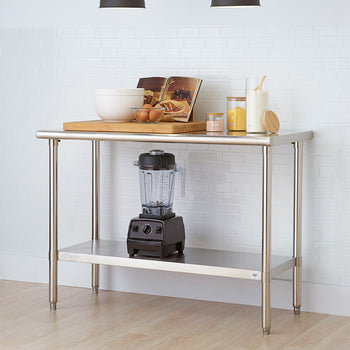A stainless steel work table with a lower shelf is setup in a brightly lit kitchen in front of a white tiled wall. The tabletop features a large wooden cutting board holding a mixing bowl, eggs, and an open cookbook, along with ingredient jars, while a blender sits on the bottom shelf.