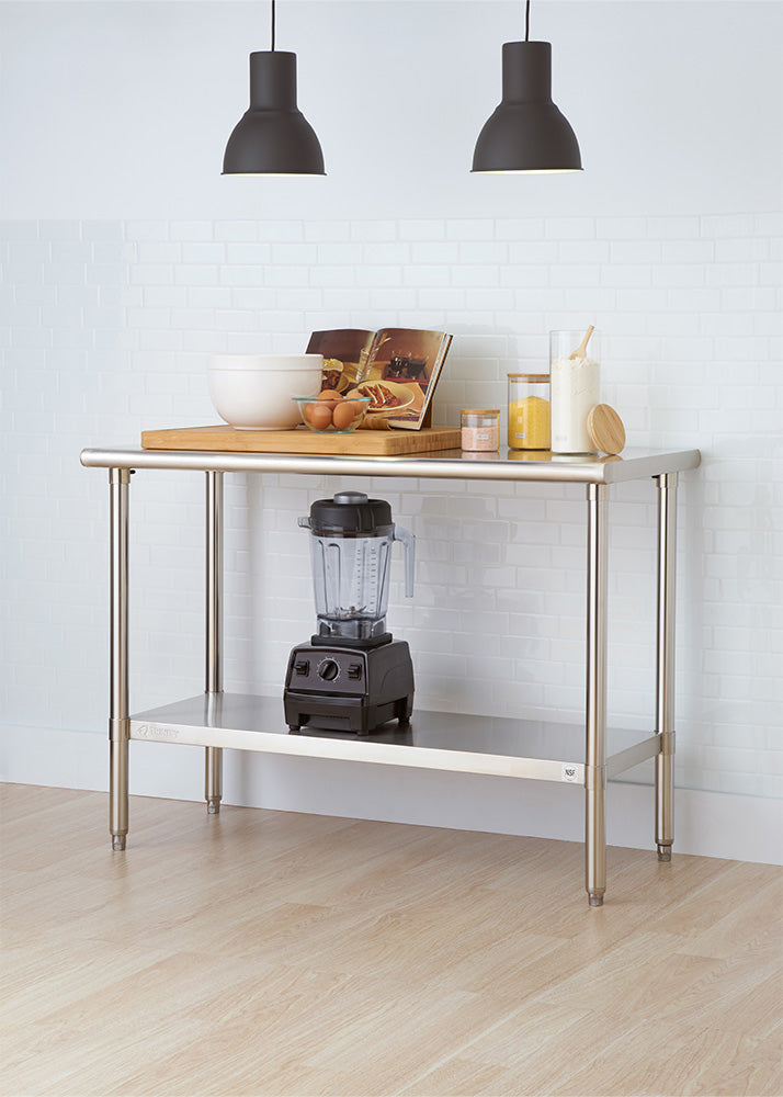 A stainless steel work table with a lower shelf is setup in a brightly lit kitchen in front of a white tiled wall. The tabletop features a large wooden cutting board holding a mixing bowl, eggs, and an open cookbook, along with ingredient jars, while a blender sits on the bottom shelf.