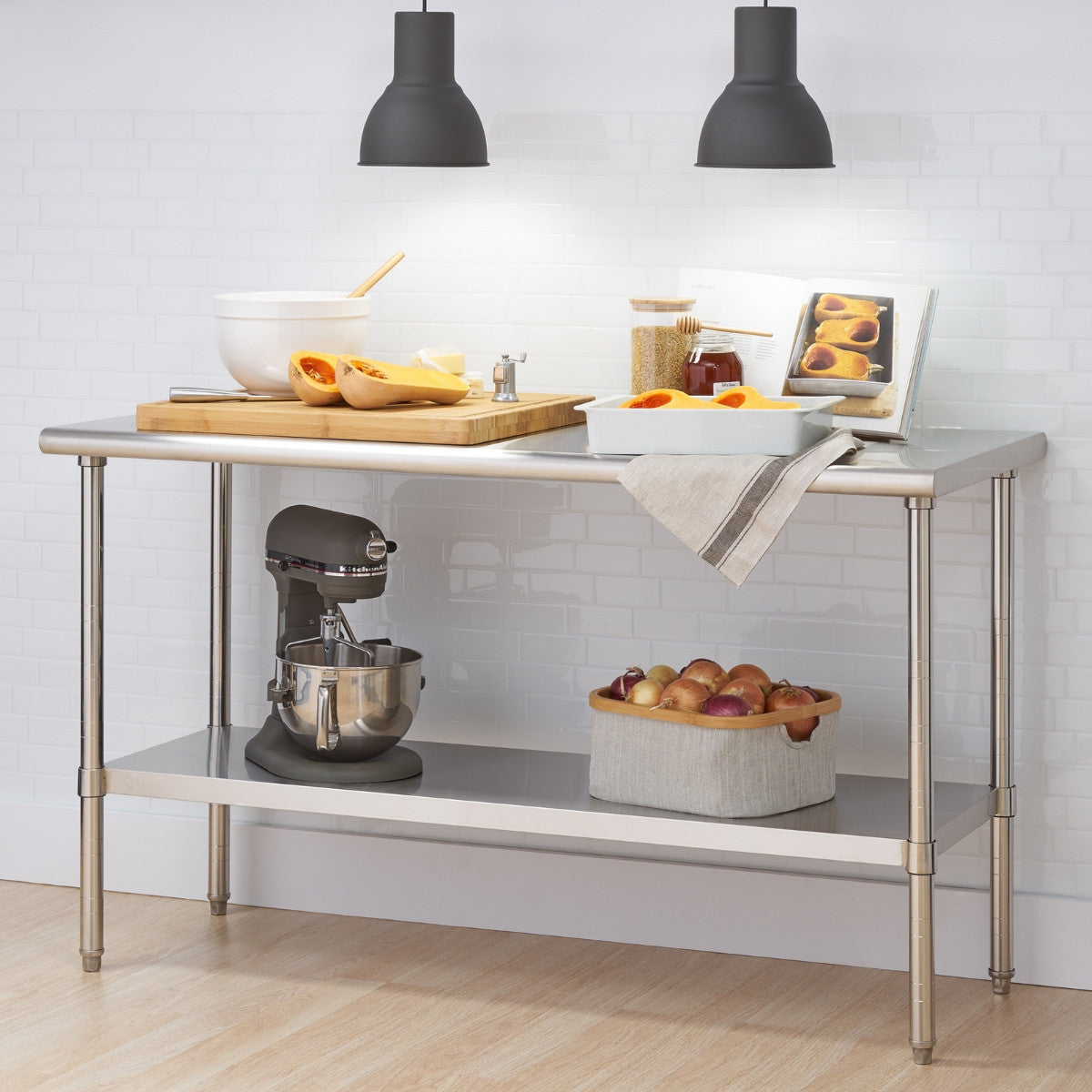 A stainless steel kitchen prep table stands against a white brick wall, illuminated by two hanging black pendant lights. The table holds ingredients for cooking, including butternut squash, a mixing bowl, a cutting board, and a cookbook, while a stand mixer and a basket of onions sit on the shelf below.