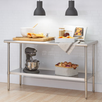 A stainless steel kitchen prep table stands against a white brick wall, illuminated by two hanging black pendant lights. The table holds ingredients for cooking, including butternut squash, a mixing bowl, a cutting board, and a cookbook, while a stand mixer and a basket of onions sit on the shelf below.