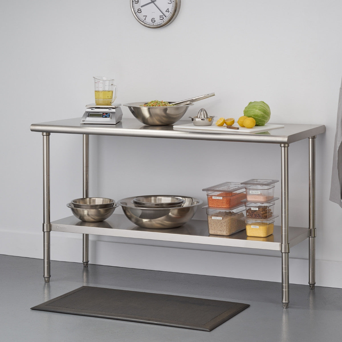 A stainless steel kitchen utility table with two shelves stands against a white wall. The top shelf holds a digital scale with a measuring cup, a large metal bowl with food and tongs, a white cutting board with lemons, a lime, and a head of lettuce, and a metal citrus juicer. The lower shelf contains several metal mixing bowls and clear plastic food storage containers filled with various ingredients.