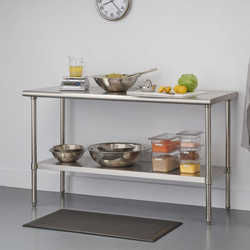 A stainless steel kitchen utility table with two shelves stands against a white wall. The top shelf holds a digital scale with a measuring cup, a large metal bowl with food and tongs, a white cutting board with lemons, a lime, and a head of lettuce, and a metal citrus juicer. The lower shelf contains several metal mixing bowls and clear plastic food storage containers filled with various ingredients.