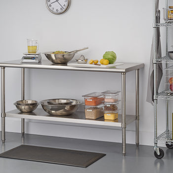 A professional stainless steel prep table stands in a clean, white kitchen. On the top shelf, ingredients like onions, lemons, and salad are ready to be used, while the lower shelf holds large mixing bowls and clear containers of dry goods.