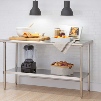 A stainless steel commercial work table features a large wooden cutting board with halved butternut squash, prep bowls, and a cookbook illuminated by two overhead black pendant lights. The bottom storage shelf conveniently holds a countertop blender and a basket filled with onions against a white subway tile backdrop.
