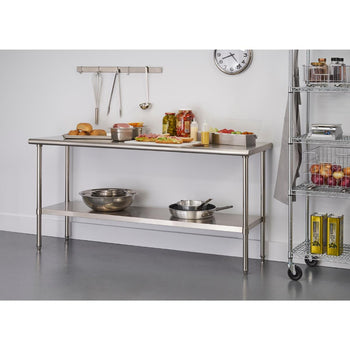 A stainless steel kitchen prep table stands in a kitchen setting. The table has two tiers and holds various food items, bowls, and a frying pan, while kitchen utensils hang on the wall behind it.