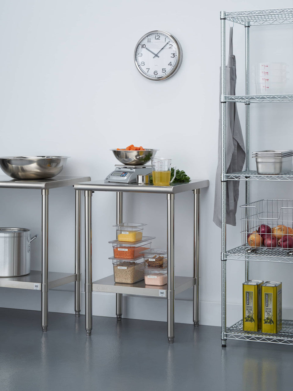 A bright kitchen space features two stainless steel utility tables. The table on the left holds a large stainless steel mixing bowl, while the table on the right has a digital scale with a bowl of carrots, a container of lentils, and other kitchen items.