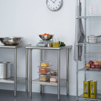 A bright kitchen space features two stainless steel utility tables. The table on the left holds a large stainless steel mixing bowl, while the table on the right has a digital scale with a bowl of carrots, a container of lentils, and other kitchen items.
