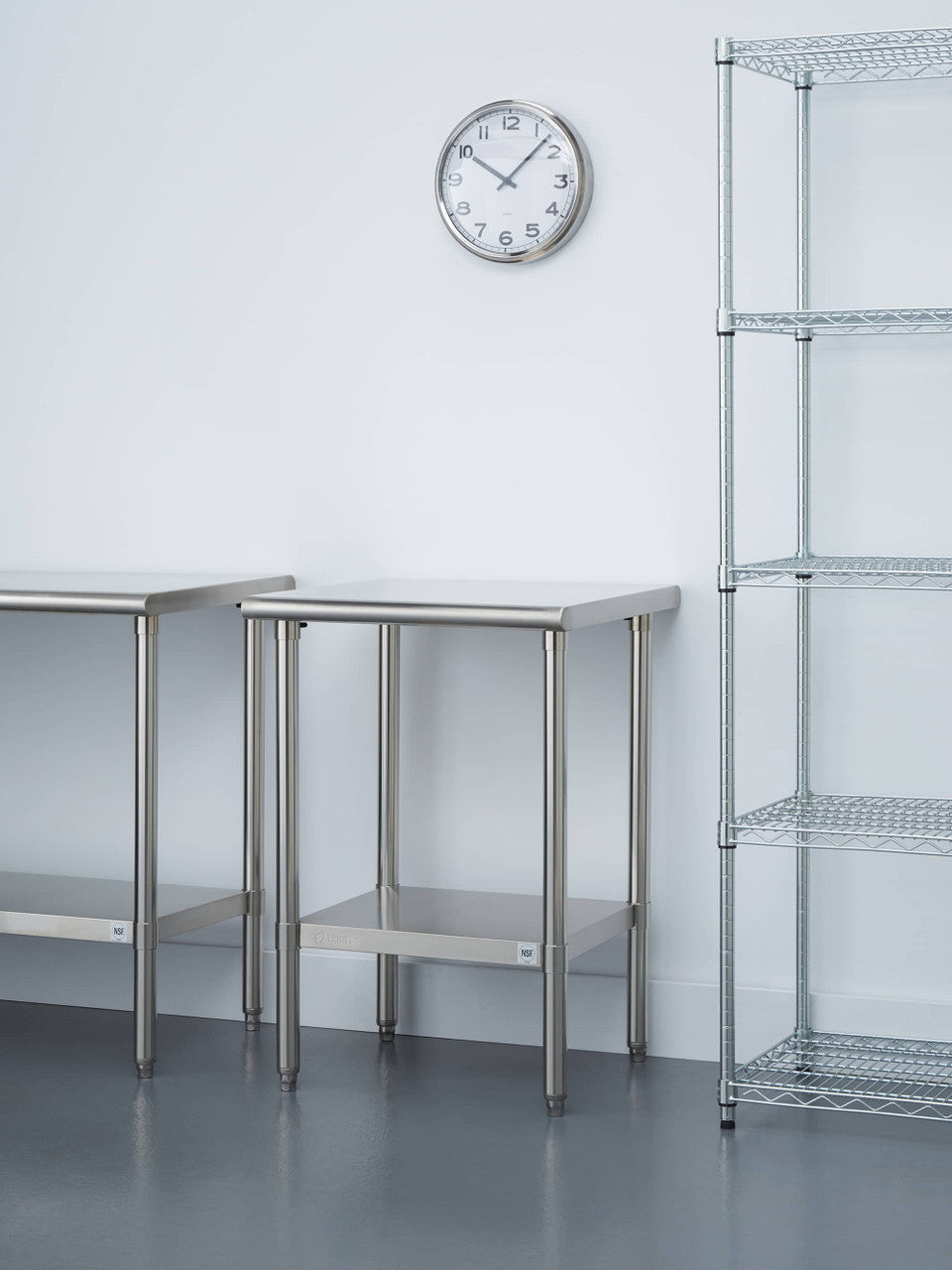 A brightly lit room features two stainless steel tables with matching lower shelves. A round analog clock hangs on the white wall above the tables, and a tall, four-tiered wire shelving unit stands to the right.