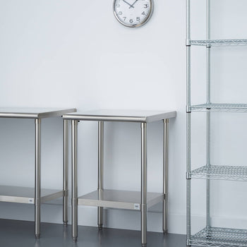 A brightly lit room features two stainless steel tables with matching lower shelves. A round analog clock hangs on the white wall above the tables, and a tall, four-tiered wire shelving unit stands to the right.