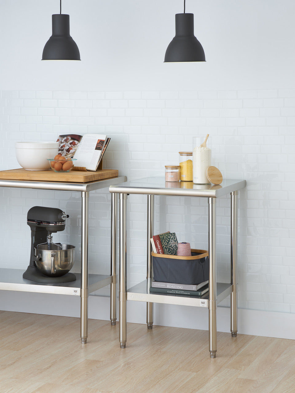 A kitchen scene features two stainless steel utility tables against a white brick wall. The left table holds a stand mixer, a wooden cutting board with a bowl, eggs, and a cookbook, while the right table has jars, a container of flour with a scoop, a basket with linens, and books.