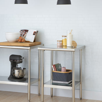 A kitchen scene features two stainless steel utility tables against a white brick wall. The left table holds a stand mixer, a wooden cutting board with a bowl, eggs, and a cookbook, while the right table has jars, a container of flour with a scoop, a basket with linens, and books.