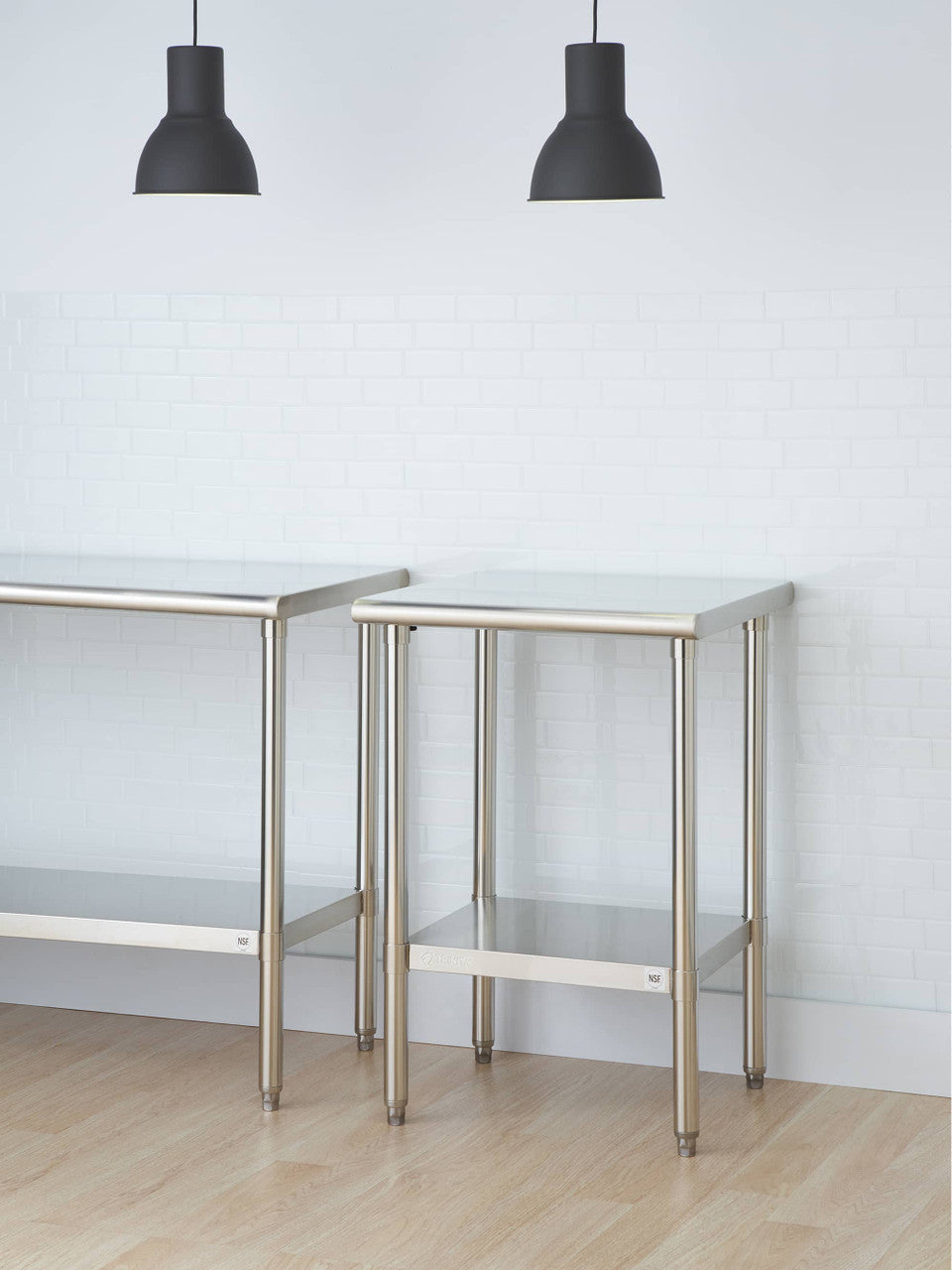 Two stainless steel tables with undershelves stand on a light wooden floor against a white brick wall. Above them, two black pendant lights hang from the ceiling.