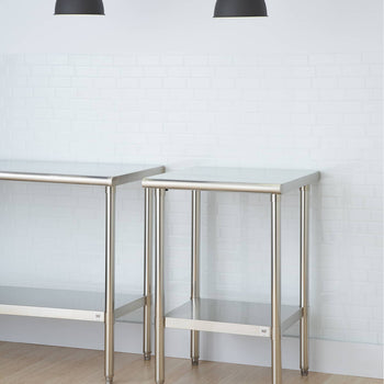 Two stainless steel tables with undershelves stand on a light wooden floor against a white brick wall. Above them, two black pendant lights hang from the ceiling.