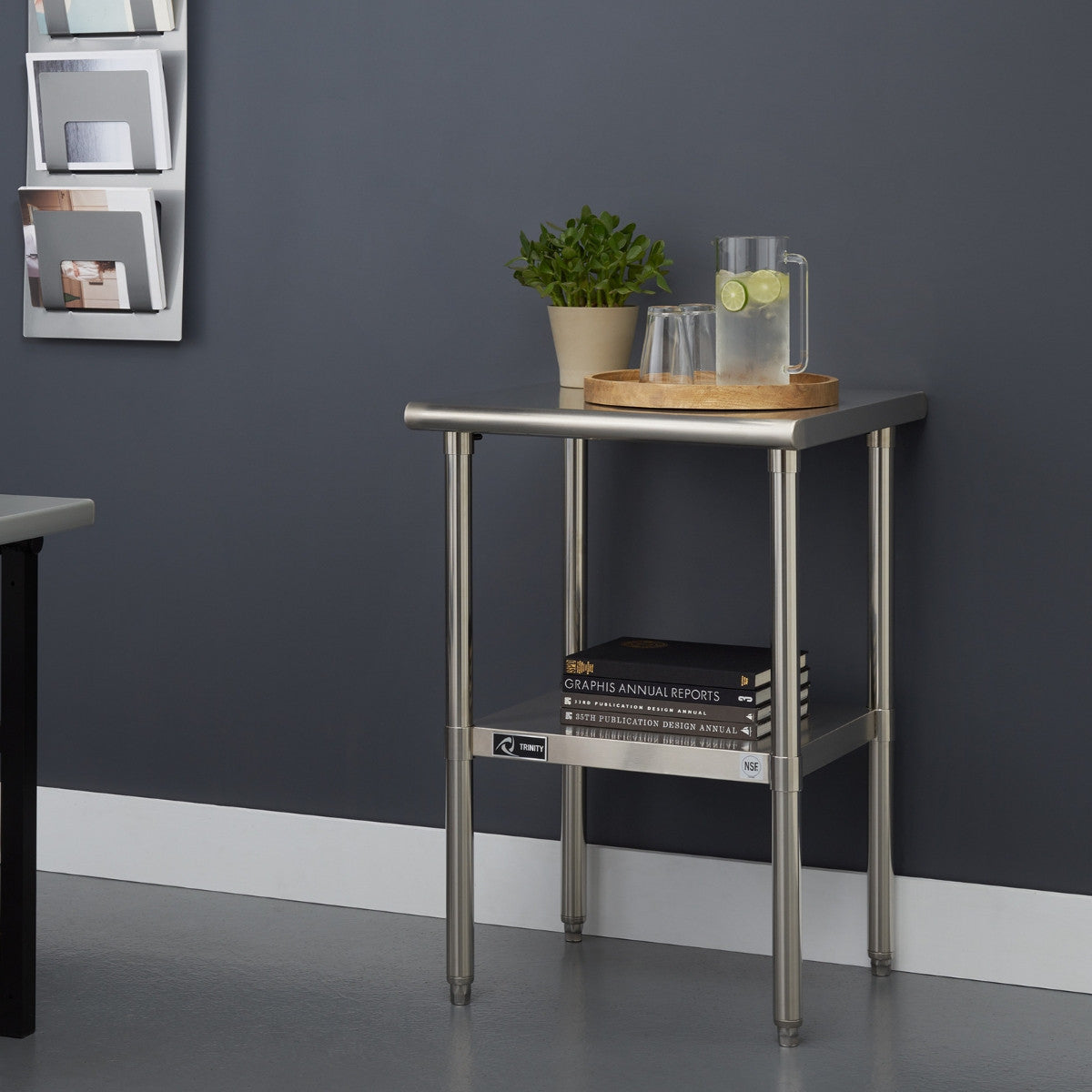 A stainless steel utility table with a square top and four cylindrical legs stands against a dark gray wall. The table holds a potted plant, a wooden tray with a glass pitcher of water and two drinking glasses, and a stack of books on a lower shelf.