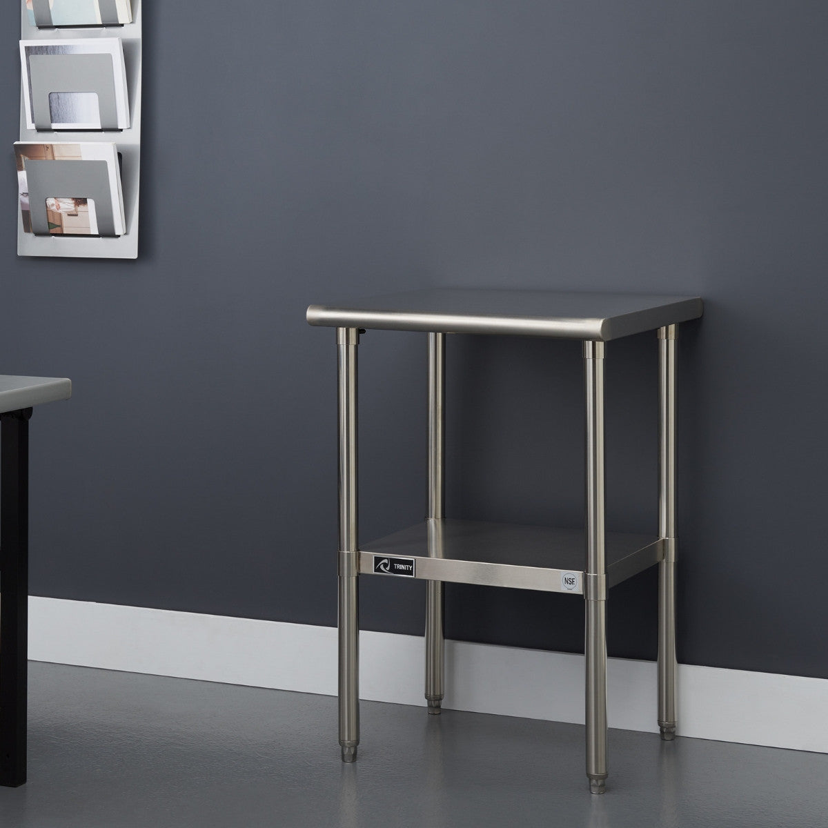 A small, square stainless steel table with four cylindrical legs and an additional shelf below stands against a dark gray wall with a white baseboard. To the left, a portion of another table with a black frame and light gray top is visible.
