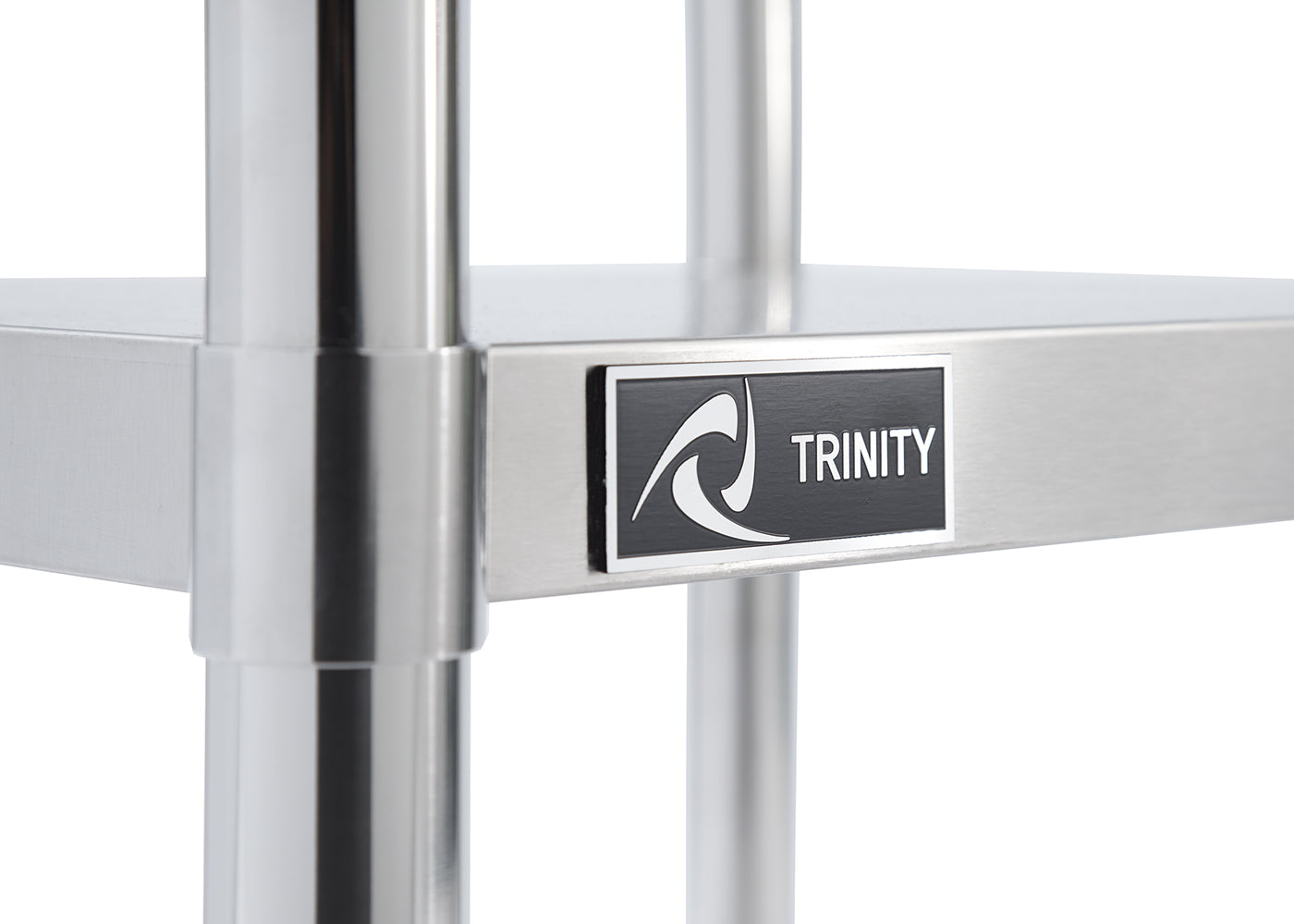 A close-up shot shows a portion of a stainless steel utility table. A black rectangular label with a white border reads "TRINITY" in white capital letters next to a stylized white wave design.