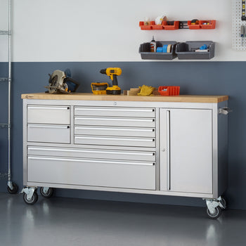 A stainless steel rolling workbench with a light wood top is shown in a garage setting. The workbench has multiple drawers on the left and a cabinet door on the right, and various tools and storage containers are placed on and above it.