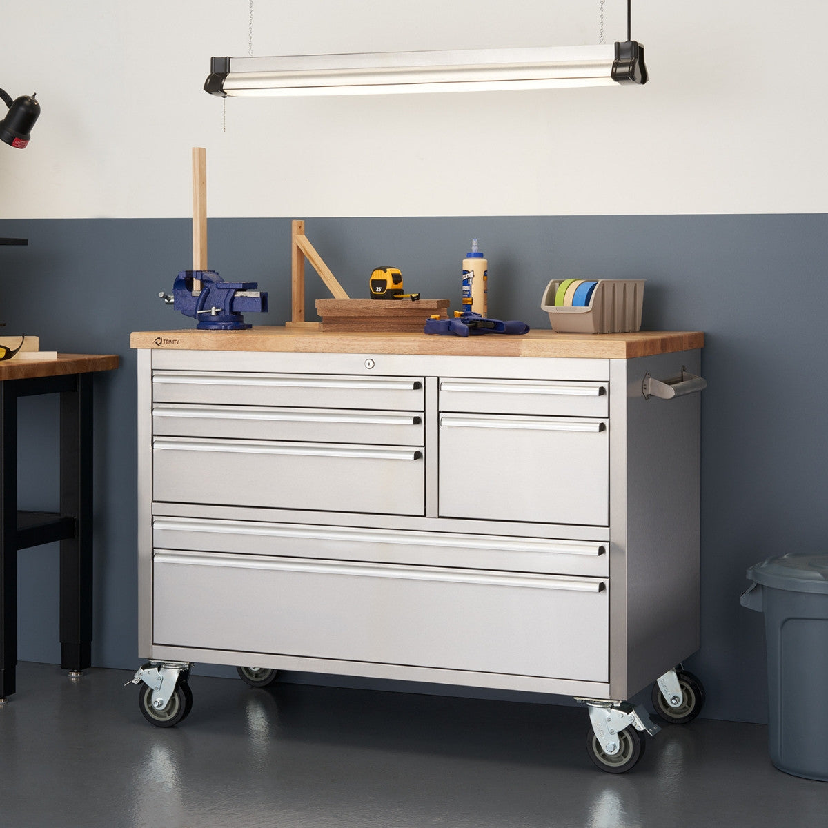 A stainless steel rolling workbench with a light-colored wooden top is positioned in a workshop setting against a two-toned wall. The workbench has multiple drawers and is equipped with wheels for mobility, and various tools and materials are arranged on its surface.