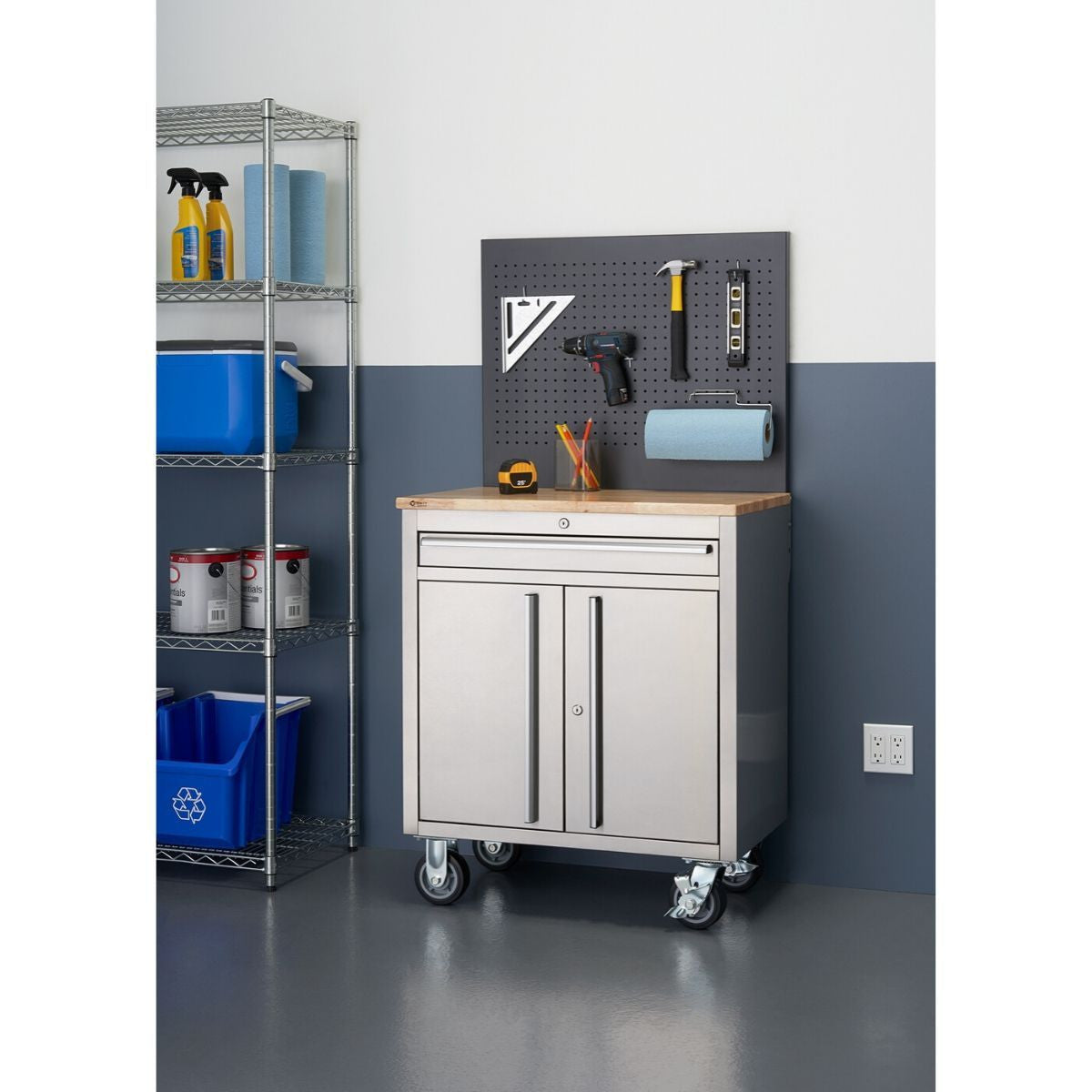 A stainless steel rolling workbench with a light wood top and a grey pegboard stands on a grey floor against a two-toned grey and white wall. Tools hang neatly on the pegboard above the workbench, which also features a drawer and a cabinet with two doors. To the left, a metal shelving unit holds various storage items.