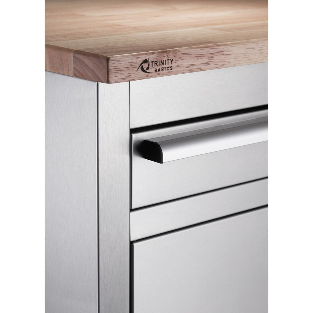 A close-up shows the corner of a Trinity Basics workbench. It features a light-colored wooden top with the "TRINITY BASICS" logo and a stainless steel body with drawer fronts and a handle visible.