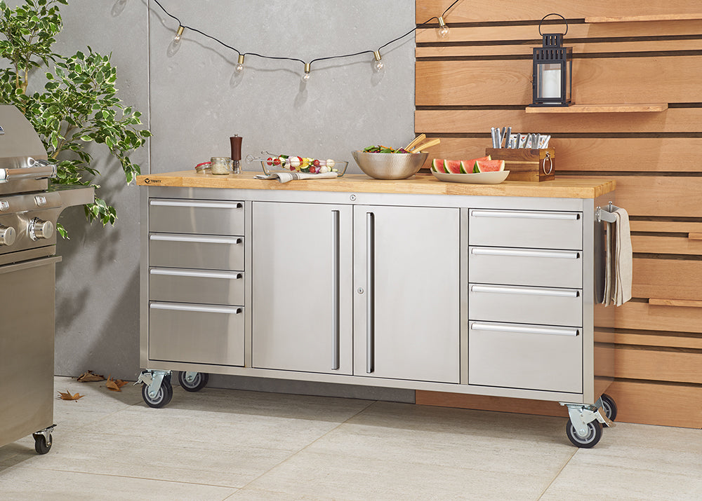 A stainless steel Trinity 72" rolling workbench with a butcher block top sits on a tiled floor, equipped with drawers and cabinets. Various food items are arranged on the wooden surface, suggesting its use as an outdoor kitchen prep station.