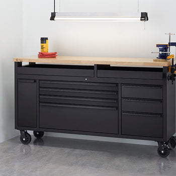 A black mobile tool workbench with a solid wood top stands fully closed in a clean workshop environment under a overhead hanging light fixture. The workstation is equipped with a bench vise and clamps attached to the side, surrounded by wire shelving, a red floor jack, and an air compressor.