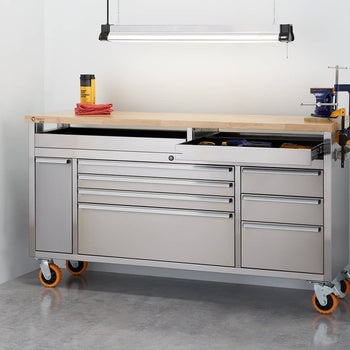 A stainless steel rolling workbench with multiple drawers and a light wood top sits in a garage setting. The workbench has orange wheels and a clamp attached to the raised section of the top.