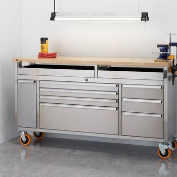 A stainless steel rolling workbench with a light-colored wooden top and multiple drawers and cabinets is positioned in a garage-like setting. The workbench has orange wheels and some tools clamped to its raised top, while other tools and equipment are visible on a nearby metal shelving unit and leaning against the wall.