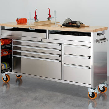 A stainless steel rolling workbench with multiple drawers and a light-colored wooden top is positioned in a workshop. The workbench has orange wheels, a raised clampable section on the top, and various tools and containers resting on it.