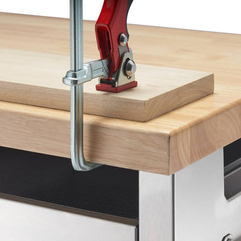 A close-up shot shows a light-colored wooden workbench with a slightly raised top edge. A red and silver clamp secures a rectangular piece of light wood to the workbench's raised edge.