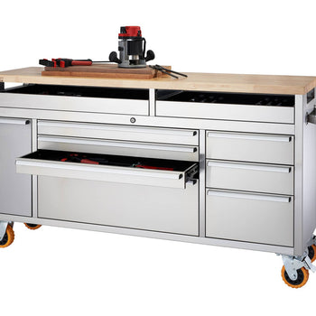 A wide shot shows a stainless steel rolling workbench with a light-colored wooden top. The workbench features multiple drawers and a cabinet, some of which are partially open revealing tools inside.