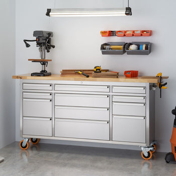 A stainless steel rolling workbench with a light wood top, various tools, and storage solutions in a workshop setting. A drill press sits on the left side of the workbench, while drawers and cabinets provide storage below, and wall-mounted organizers hold smaller items above.