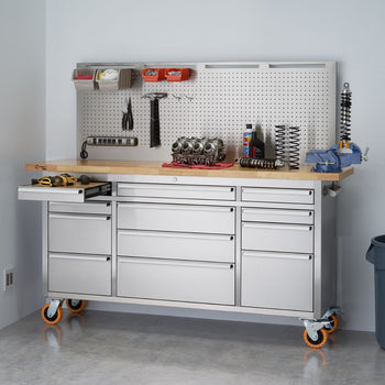 A wide shot shows a stainless steel rolling workbench with a light-colored wooden top and a pegboard backsplash against a white wall. The workbench features multiple drawers and a slide-out shelf on the left, and various tools and equipment are organized on the pegboard and the countertop, including wrenches, screwdrivers, containers, an engine part, and a vise.