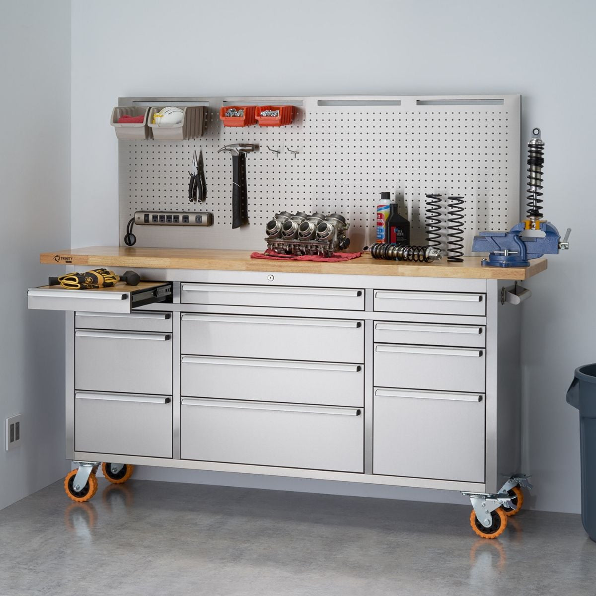 A wide shot shows a stainless steel rolling workbench with a light-colored wooden top and a pegboard backsplash against a white wall. The workbench features multiple drawers and a slide-out shelf on the left, and various tools and equipment are organized on the pegboard and the countertop, including wrenches, screwdrivers, containers, an engine part, and a vise.