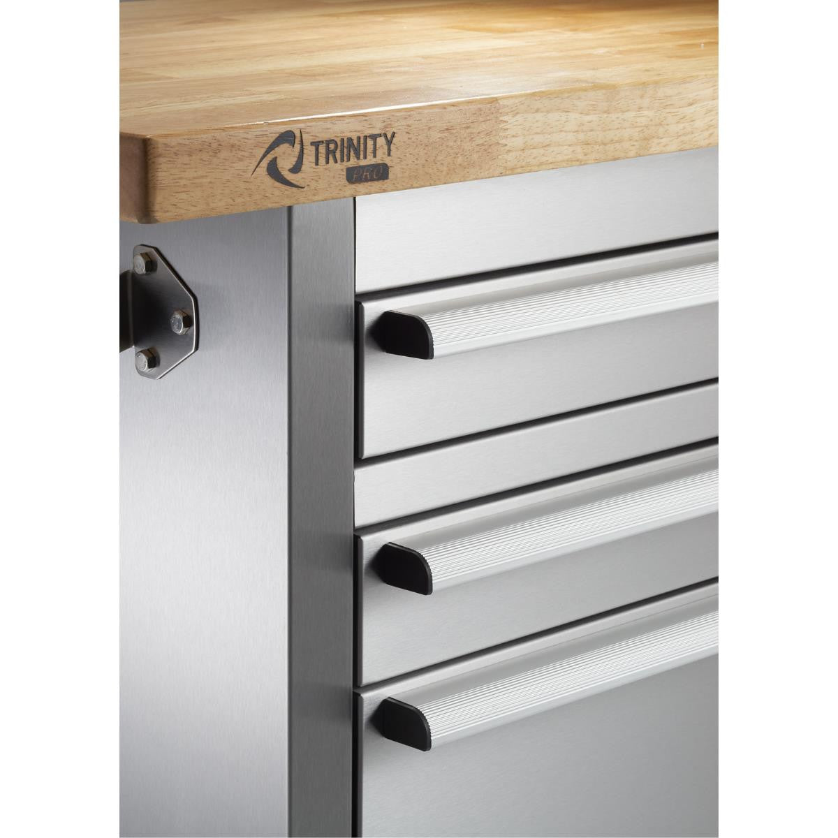 A close-up shot shows a portion of a stainless steel rolling workbench with a light-colored wooden top featuring the "TRINITY" brand logo. Three drawers with horizontal ribbed silver handles are visible on the side of the workbench.