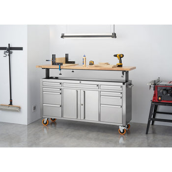 A stainless steel rolling workbench with multiple drawers and cabinets, topped with a light-colored adjustable-height wooden surface, sits in a garage or workshop. Various tools and materials are placed on the wooden top, and a fluorescent light fixture hangs above it.