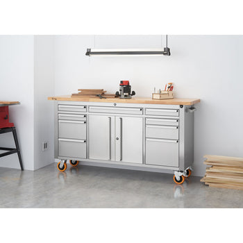 A stainless steel rolling workbench with a light wood top is positioned against a white wall in a workshop. The workbench features multiple drawers and cabinets, and rests on orange casters.