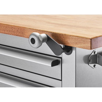 A close-up shows the adjustable mechanism of a workbench with a light-colored wooden top and stainless steel drawers. A silver handle attached to a black lever is visible on the side, suggesting a way to adjust the height or position of the workbench top.