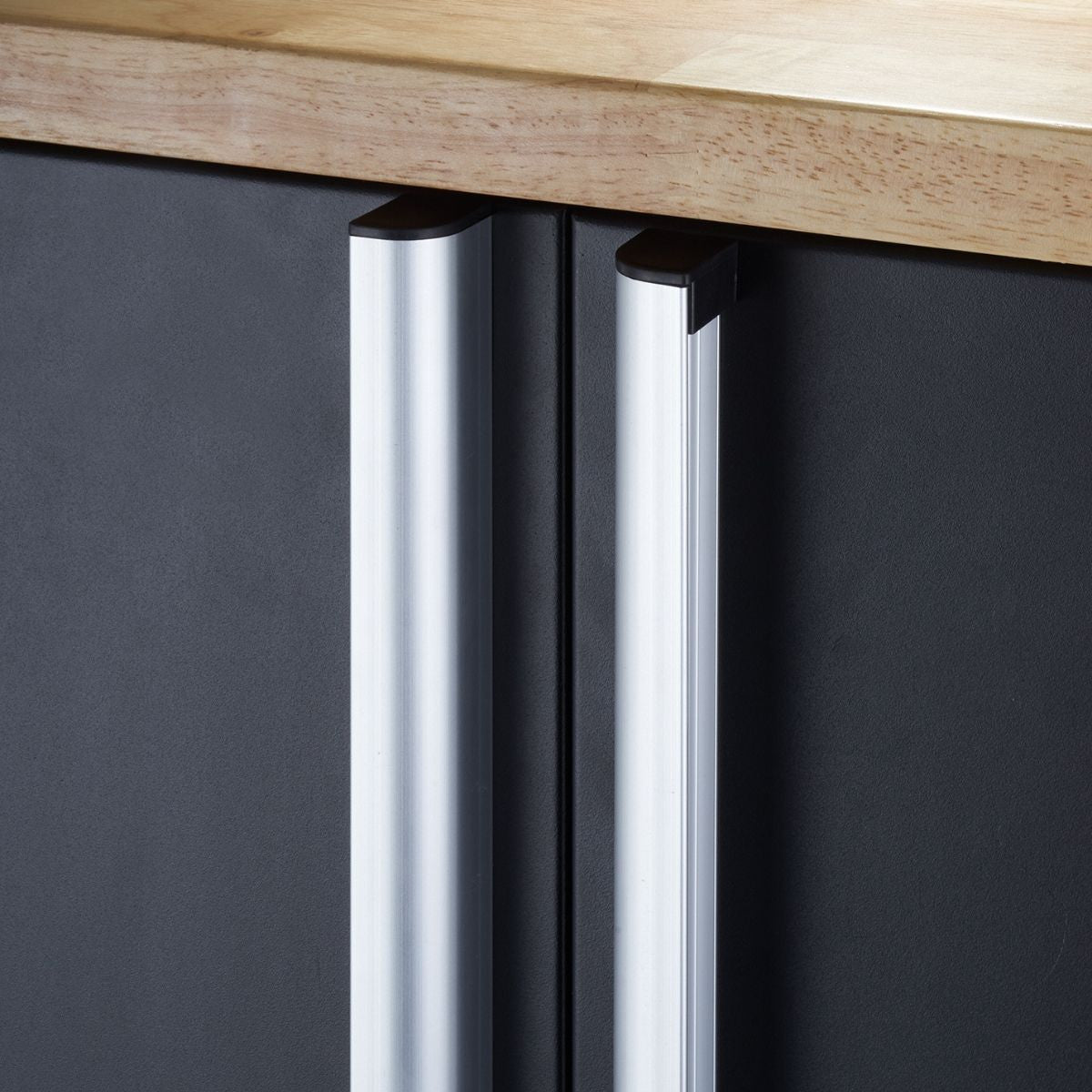 A close-up shot shows the silver handles of a black cabinet, possibly a garage base cabinet, underneath a light-colored wooden countertop. The vertical handles have a curved top and reflect light.