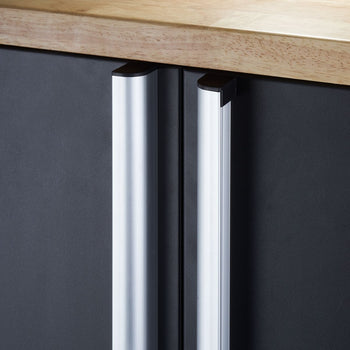 A close-up shot shows the silver handles of a black cabinet, possibly a garage base cabinet, underneath a light-colored wooden countertop. The vertical handles have a curved top and reflect light.