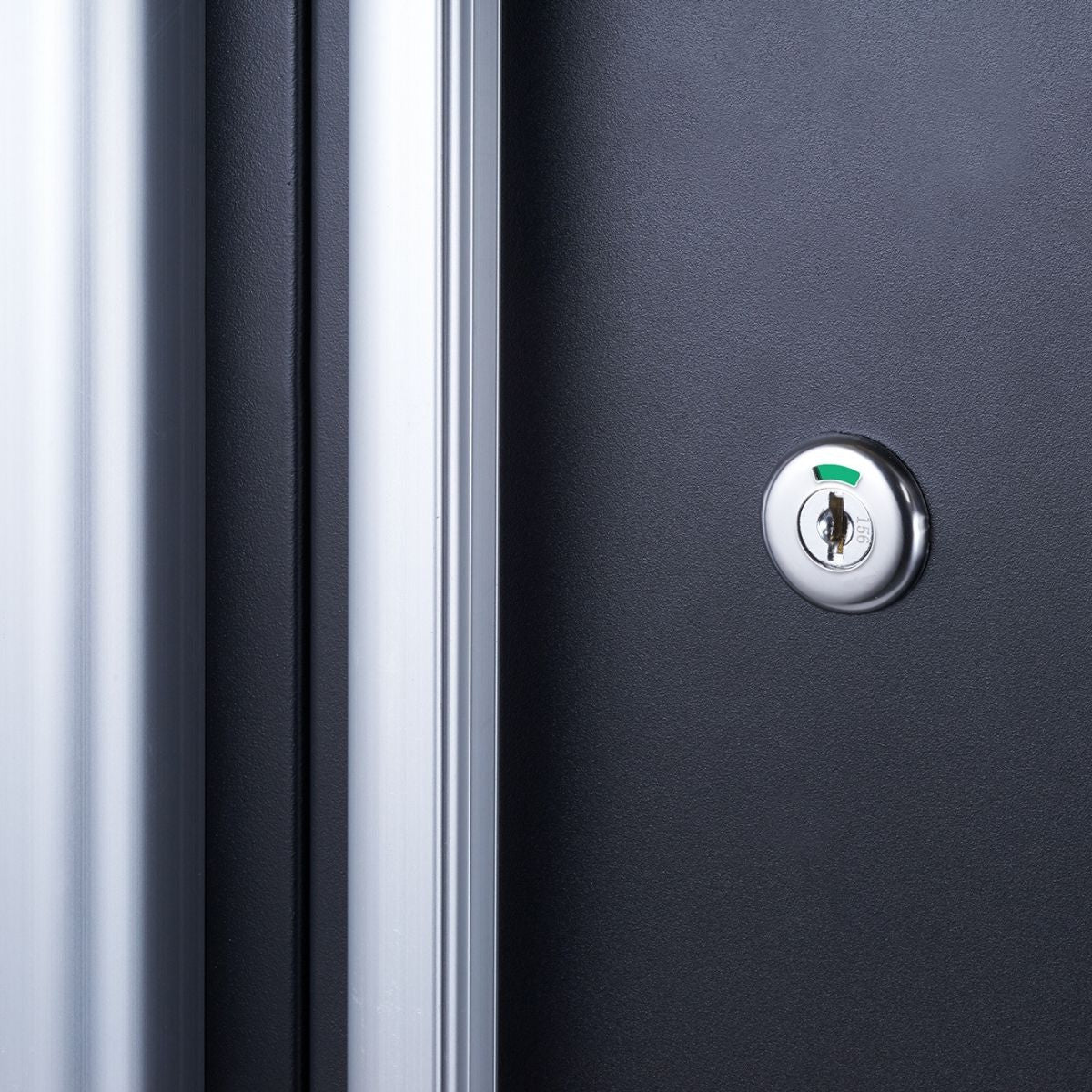 A close-up shot shows a metallic keyhole with a green indicator on a dark gray or black surface, likely a door or cabinet. To the left, a blurred vertical band of silver or light gray suggests a door frame or edge.