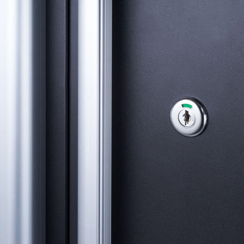 A close-up shot shows a metallic keyhole with a green indicator on a dark gray or black surface, likely a door or cabinet. To the left, a blurred vertical band of silver or light gray suggests a door frame or edge.