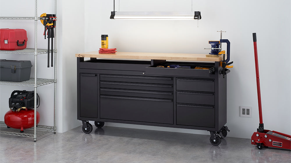 A black rolling tool workbench with a solid wooden top is positioned under an overhead light fixture in a organized garage or workshop setting. The upper right shallow drawer is pulled out, while a bench vise and clamps are mounted to the table's edge near a floor jack and wire storage shelving.