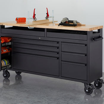 A black mobile tool workbench with a solid wood top is set up in a workshop alongside clamps, power tools, and a red air compressor. Its left-side compartment is pulled open to reveal organized storage shelves filled with orange bins and rolls of tape.