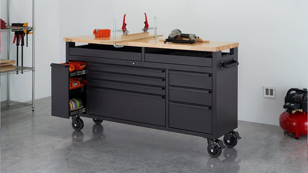 A black mobile tool workbench with a solid wood top is set up in a workshop alongside clamps, power tools, and a red air compressor. Its left-side compartment is pulled open to reveal organized storage shelves filled with orange bins and rolls of tape.
