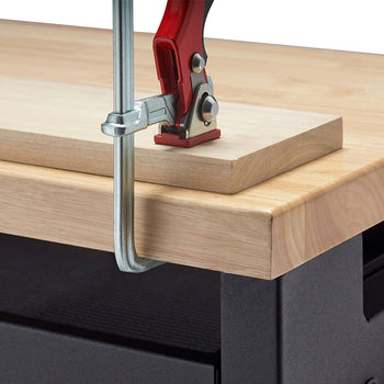 A close-up view demonstrates a heavy-duty steel bar clamp with a red handle securing a wooden plank to a workbench top. The clamp's metal arm hooks firmly around the thick butcher-block surface to ensure stability during woodworking projects.
