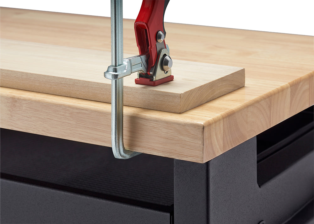 A close-up view demonstrates a heavy-duty steel bar clamp with a red handle securing a wooden plank to a workbench top. The clamp's metal arm hooks firmly around the thick butcher-block surface to ensure stability during woodworking projects.