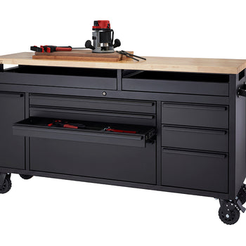 A matte black mobile tool workbench with a light wood top sits on heavy-duty locking caster wheels against a plain white background. The workbench features multiple storage drawers and side cabinets, with one drawer pulled open to reveal small hand tools while a router, clamps, and wood boards rest on the wooden surface above.